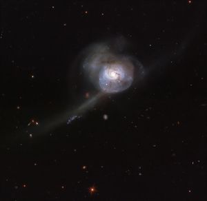 Galaxy just above center of the image with a bright white center, surrounded by pinkish-white groups of stars and brownish wisps of gas and dust, with trails of white stars off to the left. Many more galaxies in the background.